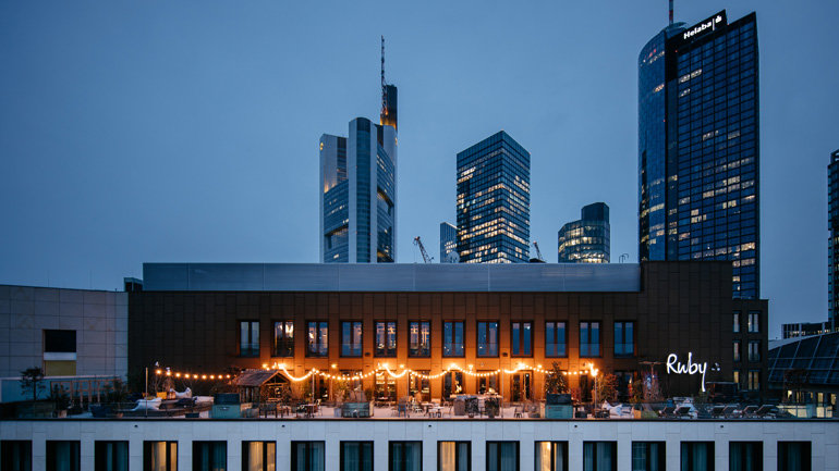 Image of Ruby Louise Hotel & Bar, Frankfurt, Germany showcasing a beautiful terrace. The image features the Ruby logo and the skyline of Frankfurt. On the terrace there is a dining area and lighting.