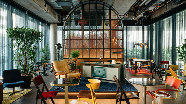 Image of Ruby Ella Hotel & Bar, Cologne, Germany showcasing the bar. The image is a classic vibe with a variety of furniture of different colours and styles. Also, including a desk laid out in the format of a chat show.