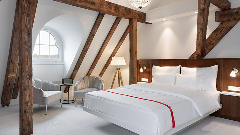 Image of Ruby Mimi Hotel & Bar, Zurich, Switzerland showcasing one of their loft style rooms. The Bedroom is broadly white with wooden features. The room features a few chairs and chandelier lighting.