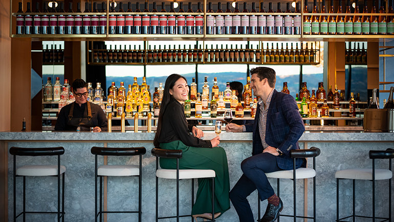 A man and a woman are sitting on two bar stool chairs at a hotel bar sipping cocktails. She is holding a glass of champagne and he is holding a glass of red wine. There is a view of mountains in the background behind the bar.