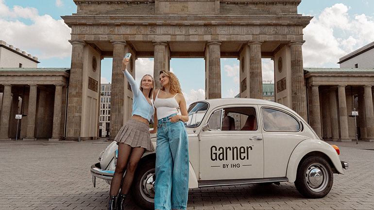 Two women taking a selfie in front of a white vintage car and the Brandenburg Gate in Berlin, Germany. The car has the "Garner BY IHG" logo on its door. The woman on the left is holding a phone up to take the photo, while the woman on the right is posing with her.