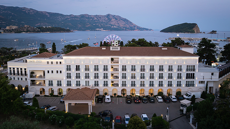An aerial shot of a large hotel complex situated on a coastline at dusk. The hotel has a multi-story, classic architectural style with many windows and balconies, illuminated by warm lights. In front of the hotel, there's a parking lot with several cars. In the background, a calm sea stretches out, dotted with boats and yachts. Further in the background, there are mountains and an island, all under a soft, twilight sky with hints of purple and blue. A large, illuminated Ferris wheel-like structure with "HB" in the center is visible on top of the hotel.