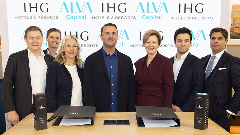 Seven business professionals pose behind a table with documents and branded items, in front of a backdrop featuring ALVA Capital, IHG Hotels & Resorts, and ALVA Hotels & Resorts logos.