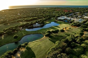 The image depicts an aerial view of Kimpton Quinta da Marinha Cascais, featuring a scenic golf course at sunset. The landscape features well-maintained green fairways, sand bunkers, and water hazards, surrounded by dense clusters of trees. In the background, you can see the resort, with the ocean visible on the horizon. The warm, golden light of the setting sun casts long shadows across the course, creating a serene and picturesque atmosphere.