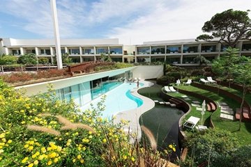 The image depicts an aerial view of Onyria Marinha Cascais, Vignette Collection. The pool is long and curvy, and people are swimming in it. The hotel buildings are white and modern, and there are comfy chairs all around the pool for relaxing. Lots of pretty flowers and green plants are growing near the pool, making it look like a secret garden. The sky is blue with white clouds, and there's a tall tree on the right side.