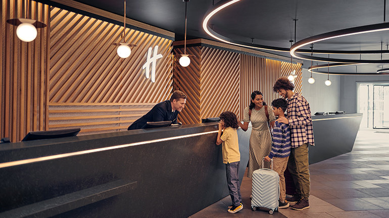Family of four checking in at a Holiday Inn hotel reception desk with luggage in a modern, contemporary lobby.