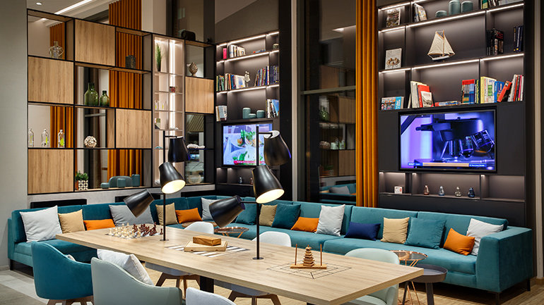 Holiday Inn open lobby featuring a large communal table with games, comfortable seating, illuminated bookshelves, and contemporary décor designed for guests to play, relax, work, and socialize.