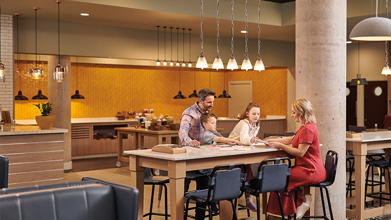 Family gathered around a communal table in a Holiday Inn open lobby, engaging in a tabletop activity within a welcoming, family-friendly shared space.