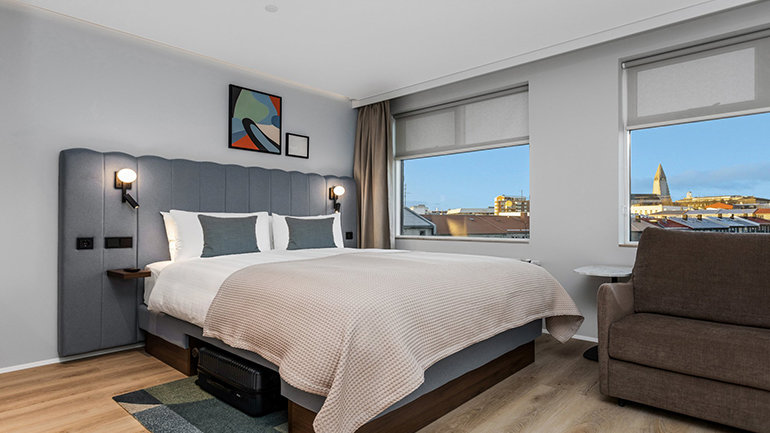 Contemporary hotel bedroom with a large double bed, neutral décor, armchair seating and windows overlooking a city skyline.