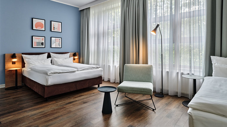 Bright hotel room with a double bed, blue feature wall, modern seating, floor lamp and large windows letting in natural light.