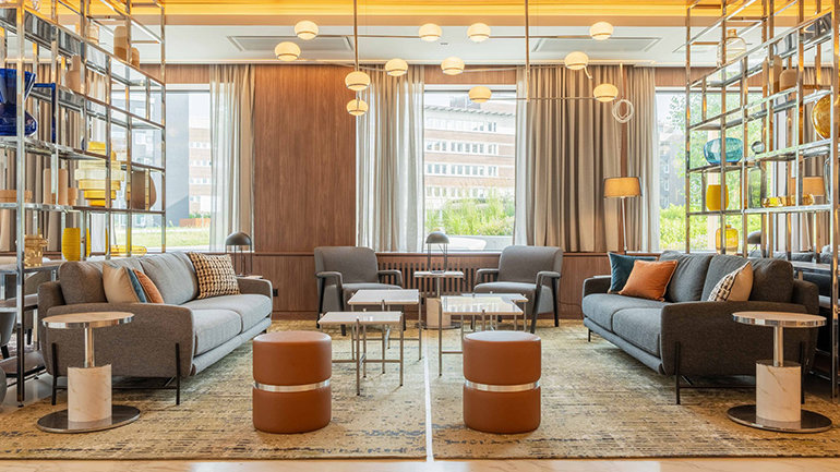Modern hotel lounge with two grey sofas facing each other, soft lighting, wooden wall panels and large windows overlooking an urban streetscape.