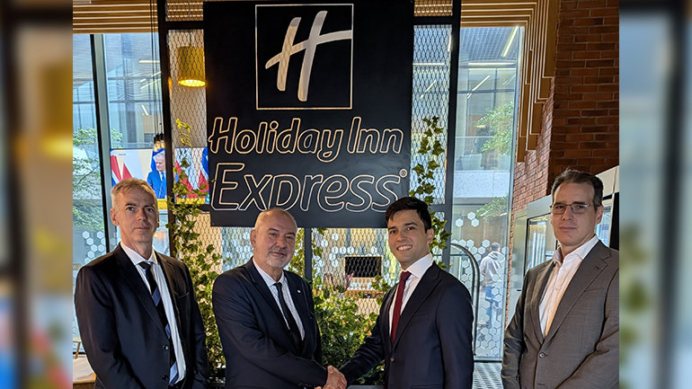 Four individuals in business attire stand inside a Holiday Inn Express hotel. Two individuals are shaking hands in front of a large Holiday Inn Express sign, with greenery and modern interior design elements in the background. The image captures a hotel development or partnership announcement.
