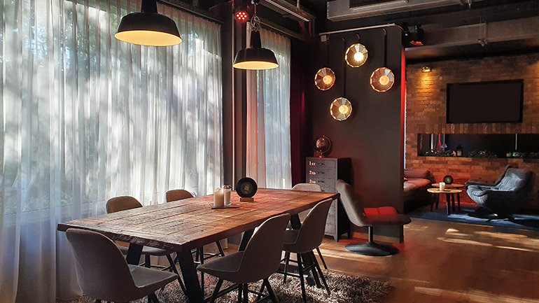Modern hotel dining and lounge space with a long wooden communal table, upholstered chairs and warm pendant lighting, set beside floor‑to‑ceiling sheer curtains and leading into a relaxed seating area with exposed brick walls.