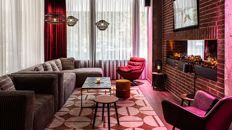 Stylish hotel lounge with plush sofas, accent chairs and a patterned rug, featuring pendant lights, a modern fireplace set into a brick wall, and floor‑to‑ceiling windows with soft curtains creating a warm, inviting atmosphere.