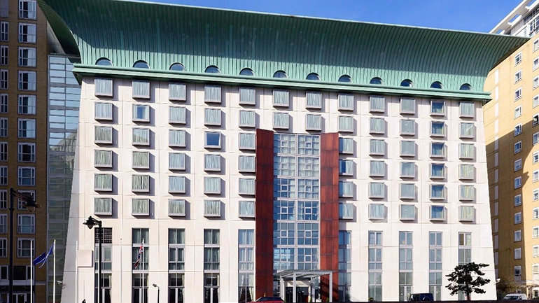 Modern hotel building with a white façade, grid of square windows and a distinctive green roof, featuring a central glass entrance framed by red panels, set between taller buildings on a clear day.