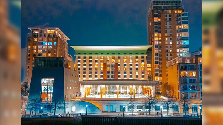 Illuminated hotel building with warm interior lights and a prominent green roof, viewed at night from across the water, surrounded by high-rise residential towers in an urban setting.