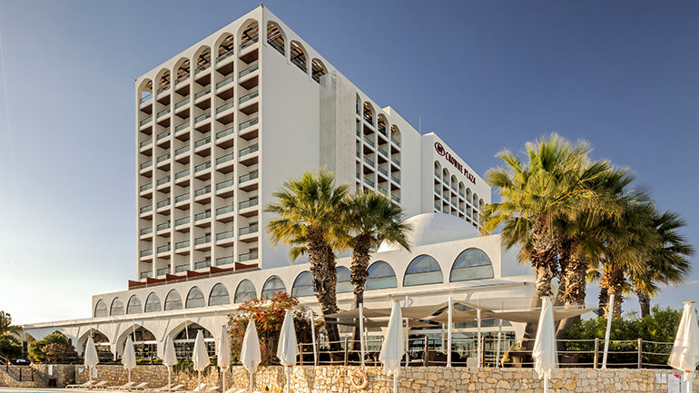 Daytime exterior shot of the current Crowne Plaza Vilamoura in Portugal, which will be converted into InterContinental Algarve – Vilamoura. The striking white multi-storey building features distinctive arched architectural detailing across its upper façade and a colonnade of arches at ground level, with the Crowne Plaza logo visible on the right-hand side. In the foreground, tall palm trees line a curved outdoor swimming pool, with white sun loungers and closed parasols arranged along a stone-clad poolside terrace. A clear blue sky completes the resort setting.