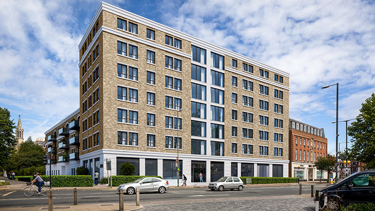 Rendering of a street-level view of a large multi-storey hotel building with a red-brick façade, white central section and rows of dark-framed windows. The building sits on a wide urban street with cars and a cyclist passing in front, trees on either side, and a blue sky with scattered clouds overhead.