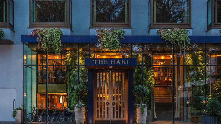 Exterior of the hotel -Street-level exterior of The Hari hotel, with its name displayed in gold lettering above a central wooden entrance. Large black-framed windows on either side reveal warmly lit interiors filled with greenery. Cascading plants and flowers hang above the windows, with potted shrubs around the entrance and several bicycles parked to the left.
