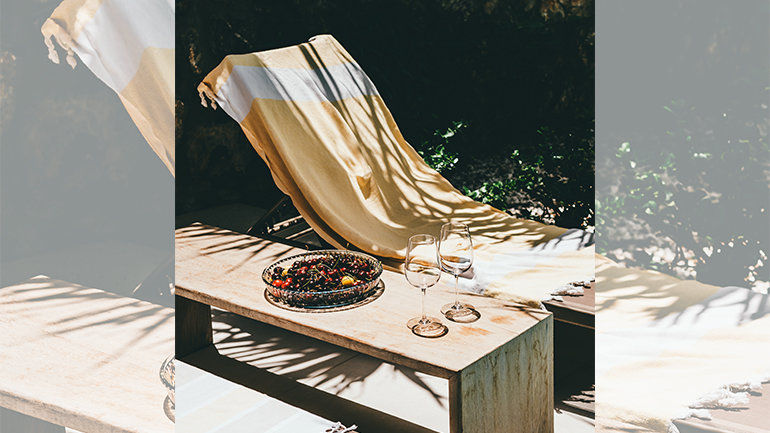 Sunlit outdoor lounge with a draped deck chair, a wooden table, two wine glasses, and a bowl of berries.