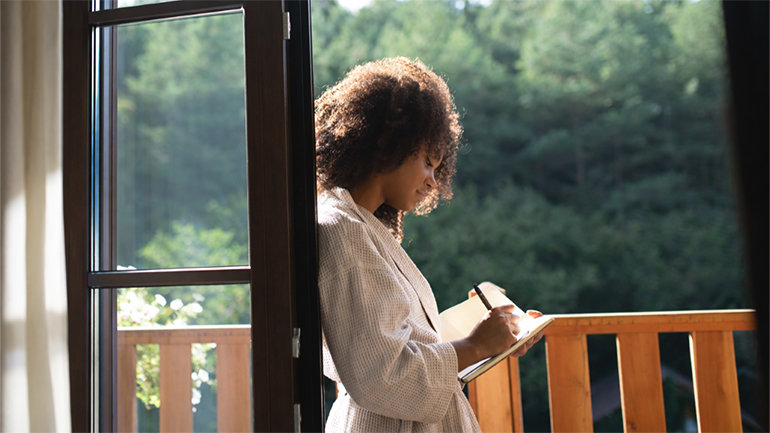Person standing by an open window, writing in a notebook while looking out over a green, wooded landscape.
