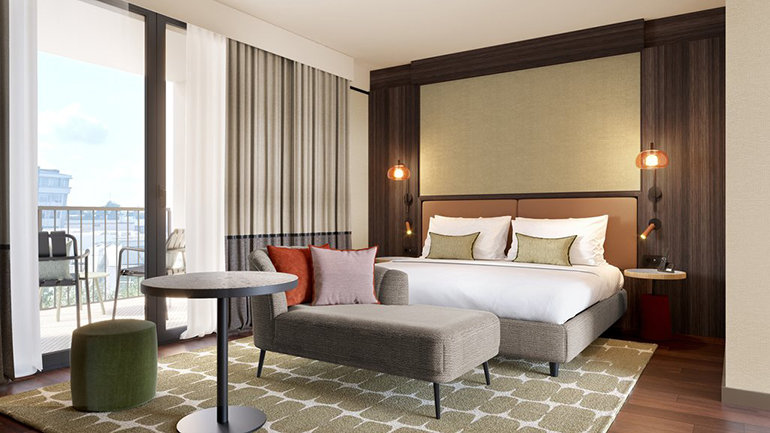 Modern hotel guest room featuring a large double bed with neutral-toned bedding, a cushioned bench at the foot of the bed, and a small seating area with a round table and upholstered chair. Floor-to-ceiling curtains open onto a balcony with outdoor seating, while warm wood panelling and soft lighting create a contemporary, inviting space.