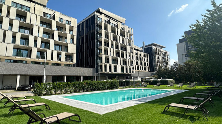 Exterior view of a contemporary hotel complex with multi-storey buildings surrounding a landscaped garden and outdoor swimming pool. Sun loungers are arranged around the pool on a well-kept lawn, with modern architecture and balconies visible under clear blue skies.