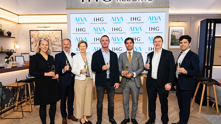 Seven people stand together holding champagne glasses at an event, in front of a backdrop displaying the IHG Hotels & Resorts and ALVA Capital logos.