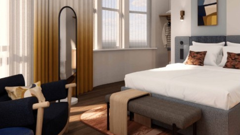 The interior of voco Bloombsury London, showing a bright, modern hotel room with large windows, a neatly made bed with accent pillows, a curved floor‑length mirror, a seating area with a navy chair, and soft curtains gradienting from white to warm gold.