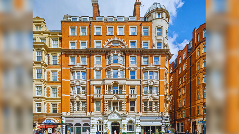 The exterior of voco Bloombsury London, showing a large, ornate red‑brick building with white stone detailing, tall windows, and a rounded corner tower, set along a busy city street with pedestrians walking by.