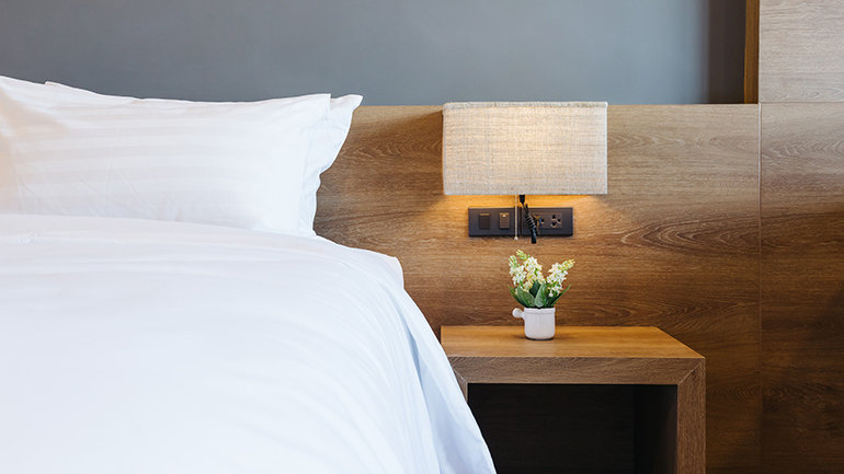 A close‑up view inside Garner Rotherham East, of a neatly made bed with crisp white linens next to a wooden bedside table, featuring a small potted plant and a textured lamp mounted on the wooden headboard.