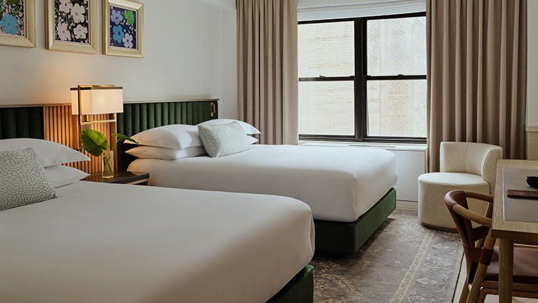 Hotel room with two queen beds featuring green upholstered headboards, white bedding, and bedside lamps, set beside a large window with beige curtains and framed artwork above the beds.