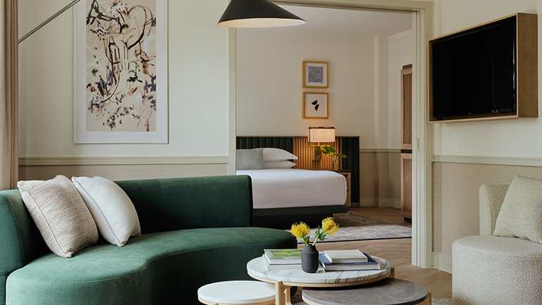 Modern hotel suite living area with a curved green sofa, layered round coffee tables, and an arched floor lamp, opening into a bedroom with a neatly made bed, abstract wall art, and soft neutral décor.
