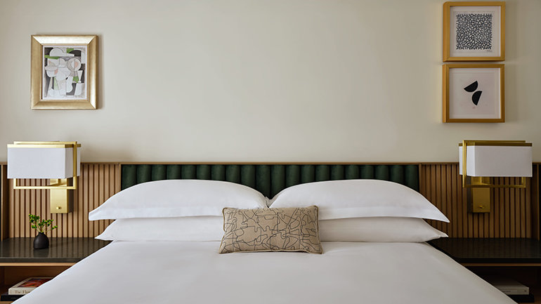 Close-up of a hotel bed with crisp white pillows and bedding, a green upholstered headboard, brass-accent bedside lamps, and minimal framed artwork on a light-colored wall.