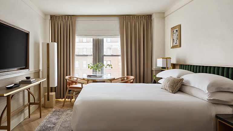 Bright hotel bedroom with a king bed dressed in white linens, green headboard, wall-mounted television, writing desk, and a small dining table near a window with floor-length curtains.