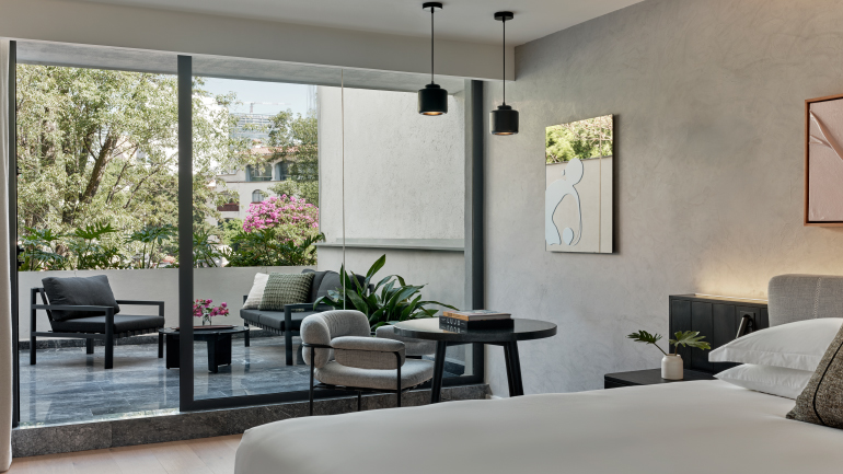 Contemporary guest room with a king bed, seating area, and floor-to-ceiling sliding glass doors opening onto a private furnished terrace surrounded by greenery. Neutral tones and modern furnishings create a bright, tranquil atmosphere.