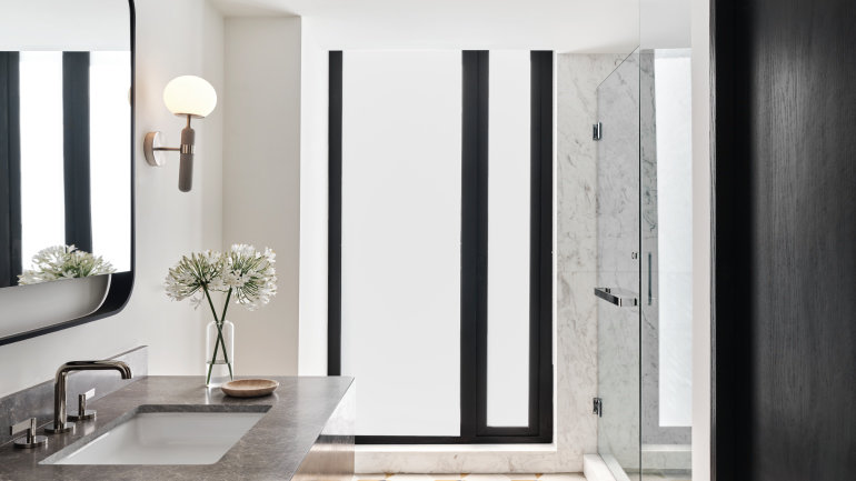 Modern bathroom featuring a gray stone vanity, rectangular sink, illuminated mirror, frosted floor-to-ceiling windows, and a glass-enclosed marble shower. A vase of white flowers adds a minimalist decorative touch.