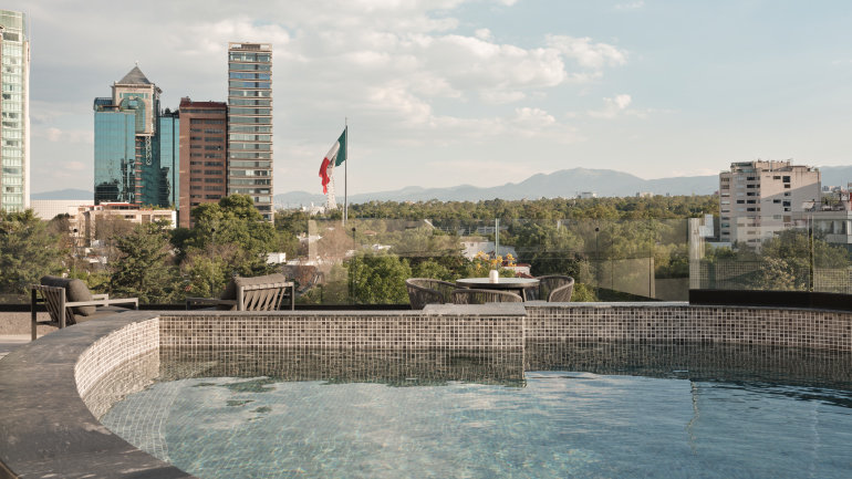 Rooftop plunge pool with glass railing overlooking a city skyline, a large Mexican flag, and distant mountains beneath a partly cloudy sky. Outdoor lounge seating and a dining table create a relaxed terrace setting.