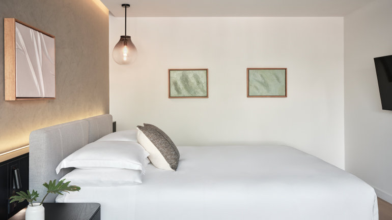 Minimalist king guest room with crisp white bedding, soft accent lighting, contemporary artwork, and a pendant light above the bedside area. Clean lines and a neutral color palette create a calm, modern retreat.