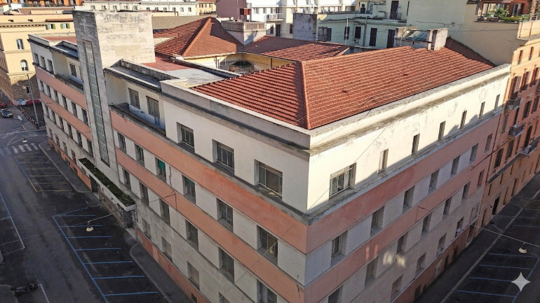 Aerial photograph of the future Kimpton Roma property on Via Sicilia in Rome, showing the existing building prior to redevelopment. The five-storey structure occupies a prominent corner site and features a cream and pale terracotta façade with a red tiled roof and a distinctive vertical glass stairwell tower. The building is surrounded by residential and mixed-use buildings in a dense urban neighbourhood, with streets and parking bays visible along both sides of the property. Taken in daylight, the image highlights the scale, footprint and city-centre location of the hotel within the surrounding Roman streetscape.