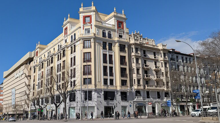Street-level exterior view of the historic six-storey building on Calle Goya in Madrid, with classical cream stone façade, red window detailing and ornate rooftop pediments — the future site of the new Kimpton Madrid hotel.