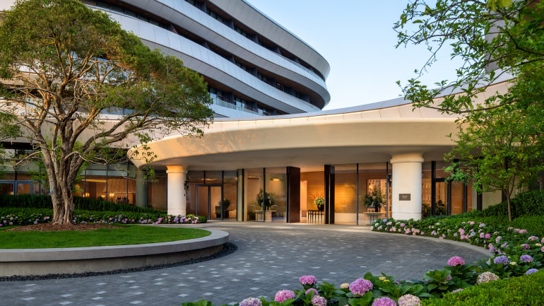 Exterior view of a contemporary luxury hotel entrance featuring a sweeping curved canopy, floor-to-ceiling glass windows, and a multi-story rounded facade with private balconies.