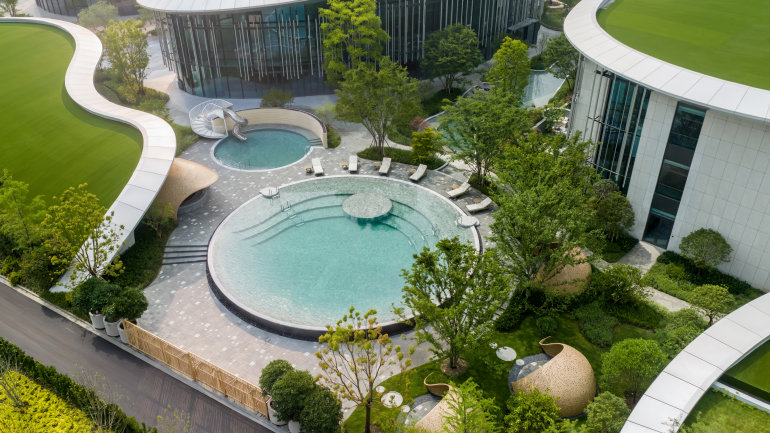 Aerial view of a modern resort courtyard featuring two circular swimming pools surrounded by lush landscaping and curved architectural buildings with green rooftop terraces. Lounge chairs line the pool decks, while winding pathways, mature trees, and sculptural garden elements create a tranquil, park-like setting between the contemporary glass-and-metal structures.