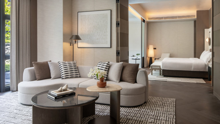 A spacious, contemporary hotel suite with a curved light-gray sofa accented by patterned and neutral-toned pillows, a black marble coffee table, and a small side table topped with white flowers.