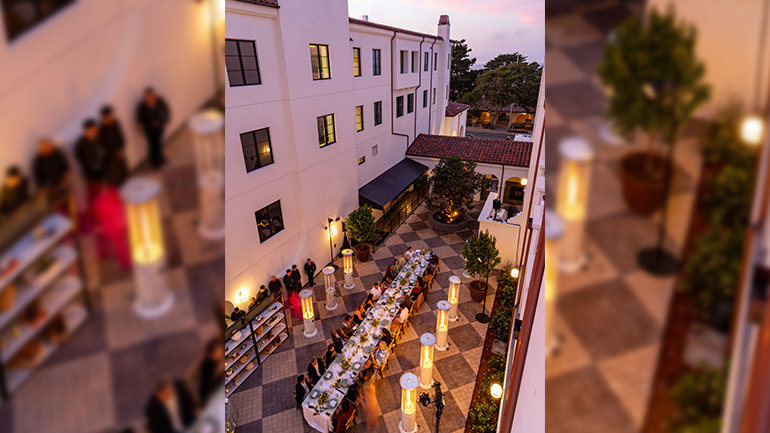 Overhead view of an outdoor courtyard dinner, with guests seated along a long, elegantly set communal table surrounded by greenery, glowing patio heaters, and white stucco buildings with red-tile roofs beneath a pink evening sky.