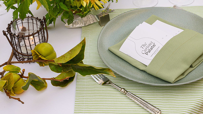Close-up of a garden-inspired table setting with a sage green plate and napkin holding a Curious Palate menu card, surrounded by sunflowers, leafy branches with green fruit, a glowing candle, glassware, and green-striped linens.