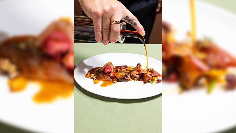 Close-up of a hand pouring a golden-brown sauce from a glass vessel over an artfully plated dish featuring thin slices, roasted vegetables, greens, edible flowers, and garnishes on a white plate.