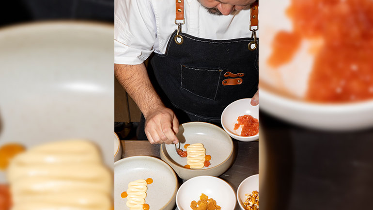 Close-up of a chef plating a dish in a kitchen, spooning a red garnish beside a piped creamy component and small dots of orange sauce in a ceramic bowl, with additional ingredients and bowls arranged nearby.