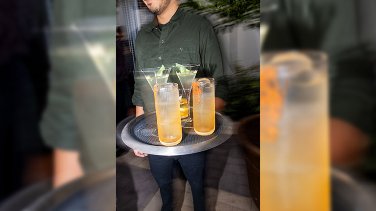 A server carries a round tray of four cocktails, including two tall golden drinks in frosted glasses and two pale cocktails in stemmed glasses garnished with green leaves.
