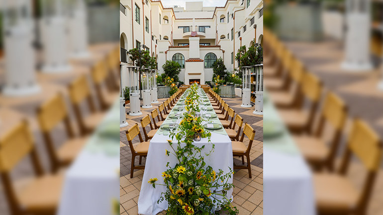 An outdoor courtyard set for a long communal dinner, with a white-clothed table lined with sage green place settings, glassware, and abundant yellow-and-white floral arrangements. Patio heaters and greenery surround the table, with a lit fireplace and white stucco building in the background.